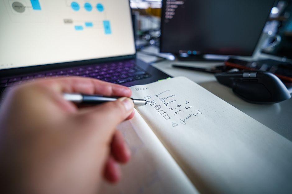 Une main prend des notes dans un carnet devant un ordinateur portable qui affiche un schéma de flux de travail