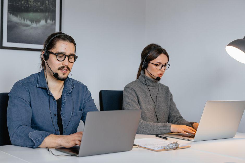 Two customer support agents wearing headsets work side by side on laptops at a white desk in a bright office
