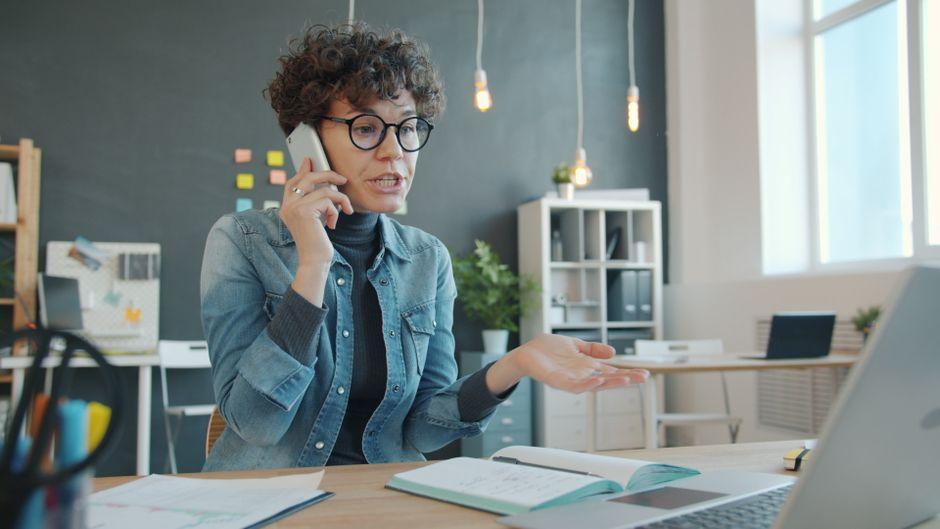 A woman in a denim jacket speaking on her phone at an office desk, with an open notebook and a laptop in front of her