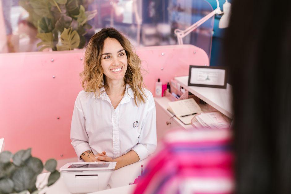 A smiling receptionist behind a pink front desk, speaking with a visitor