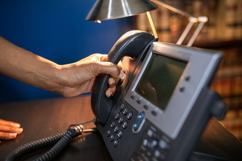 Une main saisit le combiné d'un téléphone de bureau à écran et clavier numérique, éclairé par une lampe