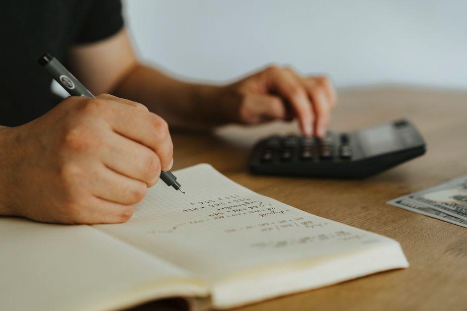 يدان تدوّنان عمليات حسابية في دفتر بجانب آلة حاسبة على مكتب خشبي