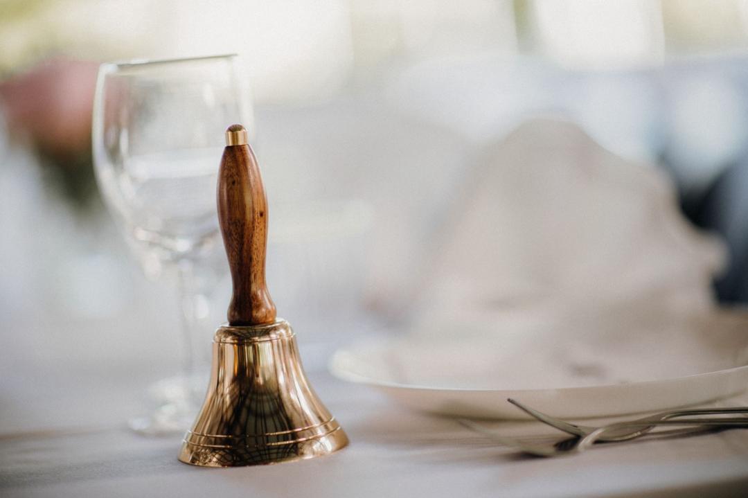 Brass hand bell with a wooden handle on a set table, with a glass and cutlery blurred behind