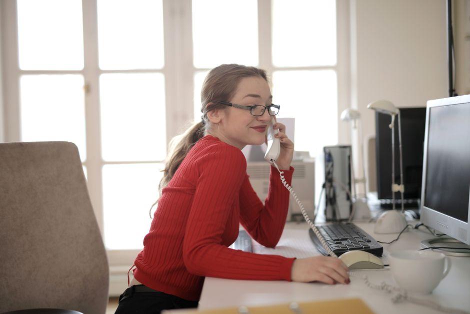 Une femme en pull rouge, lunettes sur le nez, répond au téléphone fixe à son bureau, devant une grande fenêtre, un écran d'ordinateur et une tasse à côté d'elle