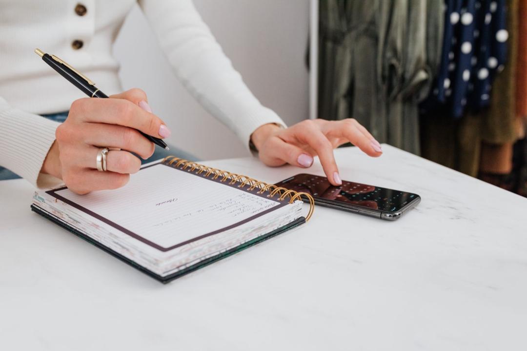 Una mano anota observaciones en un cuaderno de espiral mientras la otra usa un smartphone, sobre una mesa de mármol blanco